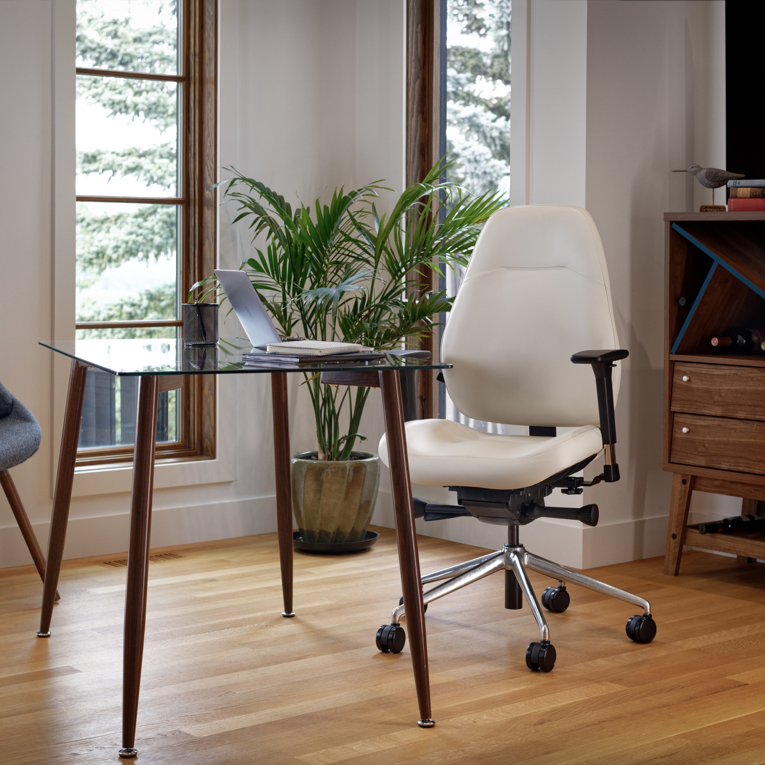 A cream-coloured ergonomic office chair with a polished chrome base sits at a glass-topped desk in a bright, modern home office with hardwood floors.