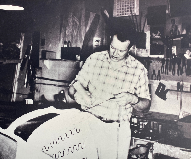 A vintage black and white photograph of a craftsman in a workshop, meticulously hand-tying the serpentine springs of a chair frame. He is focused on his task, surrounded by the tools of the upholstery trade, highlighting the hands-on skill involved in early furniture making.