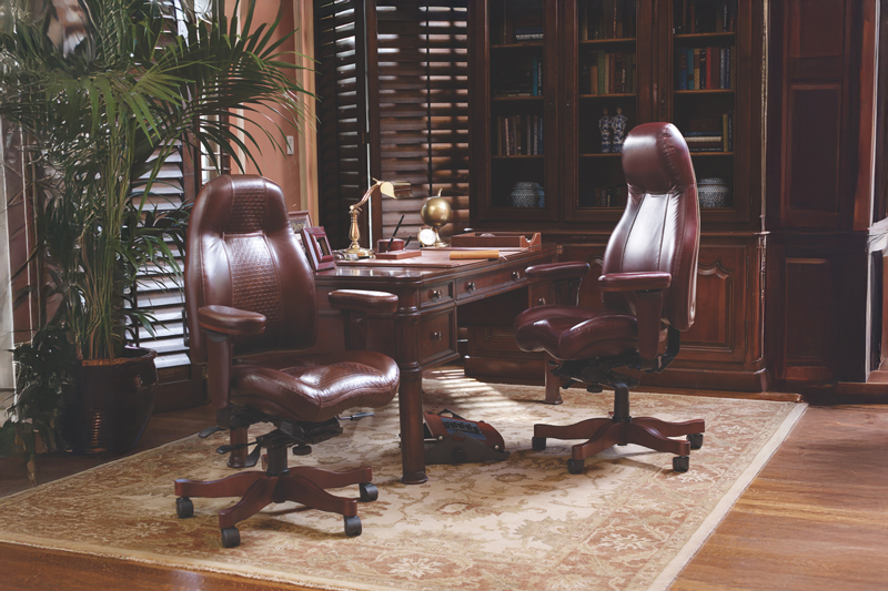 Two luxurious brown leather executive chairs, one high-back and one mid-back, are showcased in a classic, elegant office setting. The room features a traditional wooden desk, a large bookshelf filled with books, and a patterned rug, with warm sunlight streaming through the window shutters.