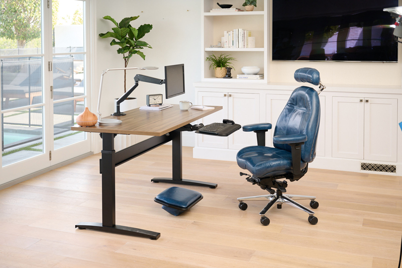 Modern home office setup featuring a height-adjustable wooden desk with a mounted monitor arm, keyboard tray, and accessories including a lamp, clock, mug, and essential oil diffuser. A luxurious blue ergonomic leather office chair with adjustable armrests and headrest sits beside the desk, with a matching footrest underneath, all positioned in a bright room with built-in shelves, a large TV, and plenty of natural light from glass doors.