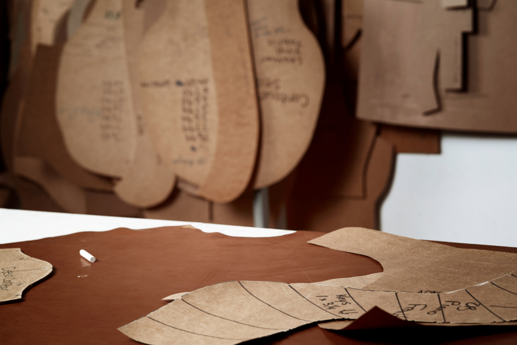A close-up of a craftsman's workbench showing paper patterns laid out on a large sheet of brown leather. A piece of white chalk rests on the leather, ready for tracing, while more patterns for chair components hang in the background of the workshop.