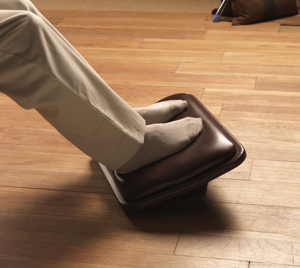 A person rests their socked feet on a cushioned, dark brown ergonomic Lifeform Chairs Executive footrest on a wooden floor.